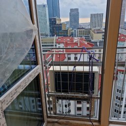 PROKLIM Łukasz Śliwiński - Montaż klimatyzatora na balkonie z widokiem na panoramę miasta. Urządzenie zabezpieczone linami, widoczne narzędzia. Okna marki Schüco.