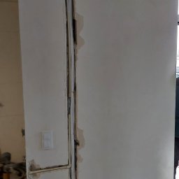 PROKLIM Łukasz Śliwiński - Biała ściana z wyżłobionym pionowym kanałem, w którym umieszczono rury i przewody. Widoczne gniazdka i włącznik. Etap prac instalacyjnych przed wykończeniem.