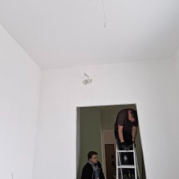 PROKLIM Łukasz Śliwiński - Montaż instalacji elektrycznej: elektryk na drabinie, w tle drugi pracownik. Przewody wystające z sufitu. Świeżo pomalowane ściany w jasnym pokoju.