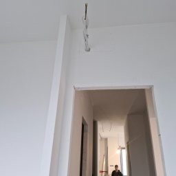 PROKLIM Łukasz Śliwiński - Wnętrze w trakcie remontu: białe ściany, wystające przewody elektryczne i sylwetka mężczyzny w oddali, sugerujące etap instalacji elektrycznej.