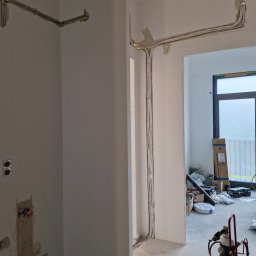 PROKLIM Łukasz Śliwiński - Instalacja przewodów wentylacyjnych/klimatyzacyjnych w surowym wnętrzu. Widoczne przewody przytwierdzone do ściany, narzędzia pracy w tle.