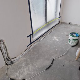 PROKLIM Łukasz Śliwiński - Instalacja przewodów w ścianie i na podłodze podczas remontu. Okno zabezpieczone folią. Widoczne kable, puszka z farbą i fragment drabiny.