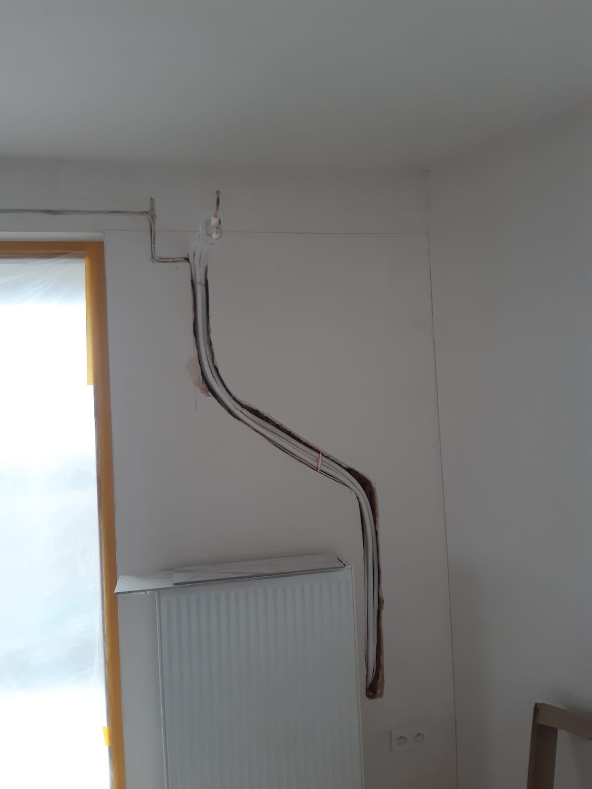 Biały grzejnik ścienny podłączony do instalacji w ścianie, widoczne przewody. Montaż systemu grzewczego w trakcie prac wykończeniowych, wnętrze pomieszczenia.