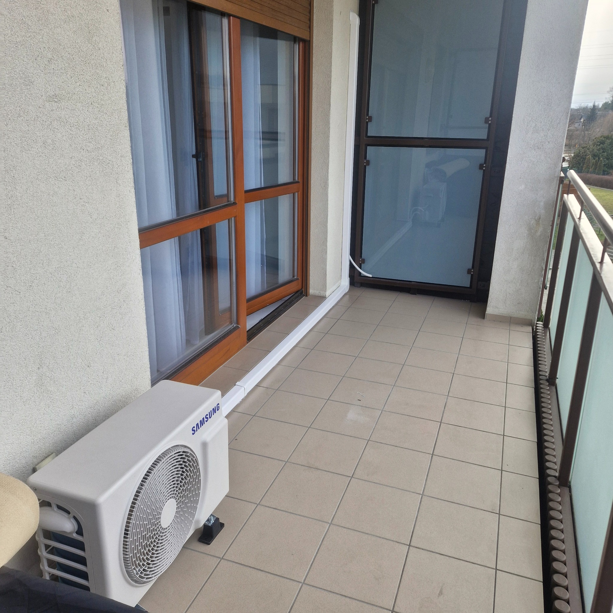 Biała jednostka zewnętrzna klimatyzacji Samsung na balkonie z brązowymi oknami i szarą podłogą, rury w listwach przypodłogowych. Widok na zewnątrz.
