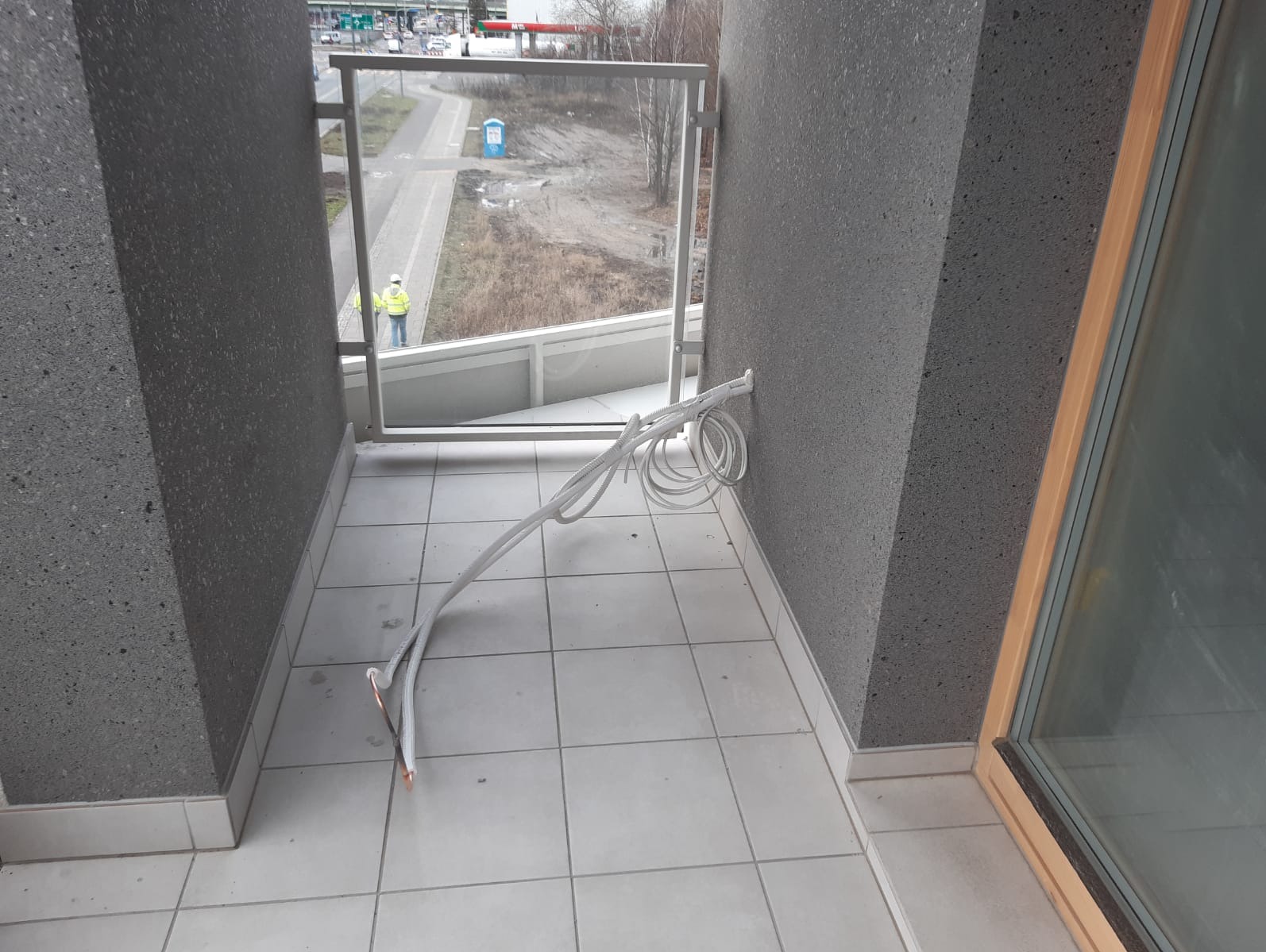 Białe kable klimatyzacji na balkonie z szarą, teksturowaną ścianą. Na zewnątrz widoczny pracownik w kamizelce. Kable częściowo zwisają.