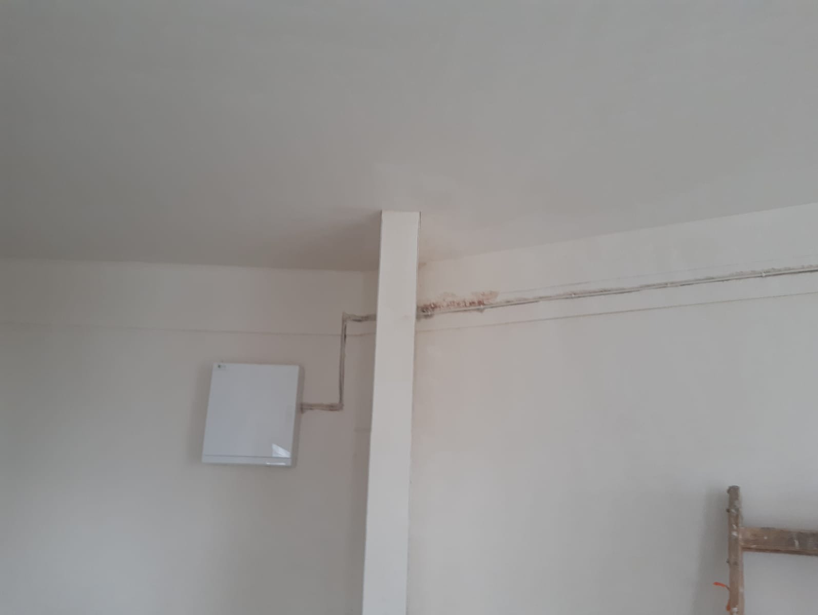 Biały rekuperator ścienny z instalacją, widok z dołu, częściowo widoczna drabina malarska. Ściany w trakcie prac wykończeniowych, widoczne ślady po instalacjach.