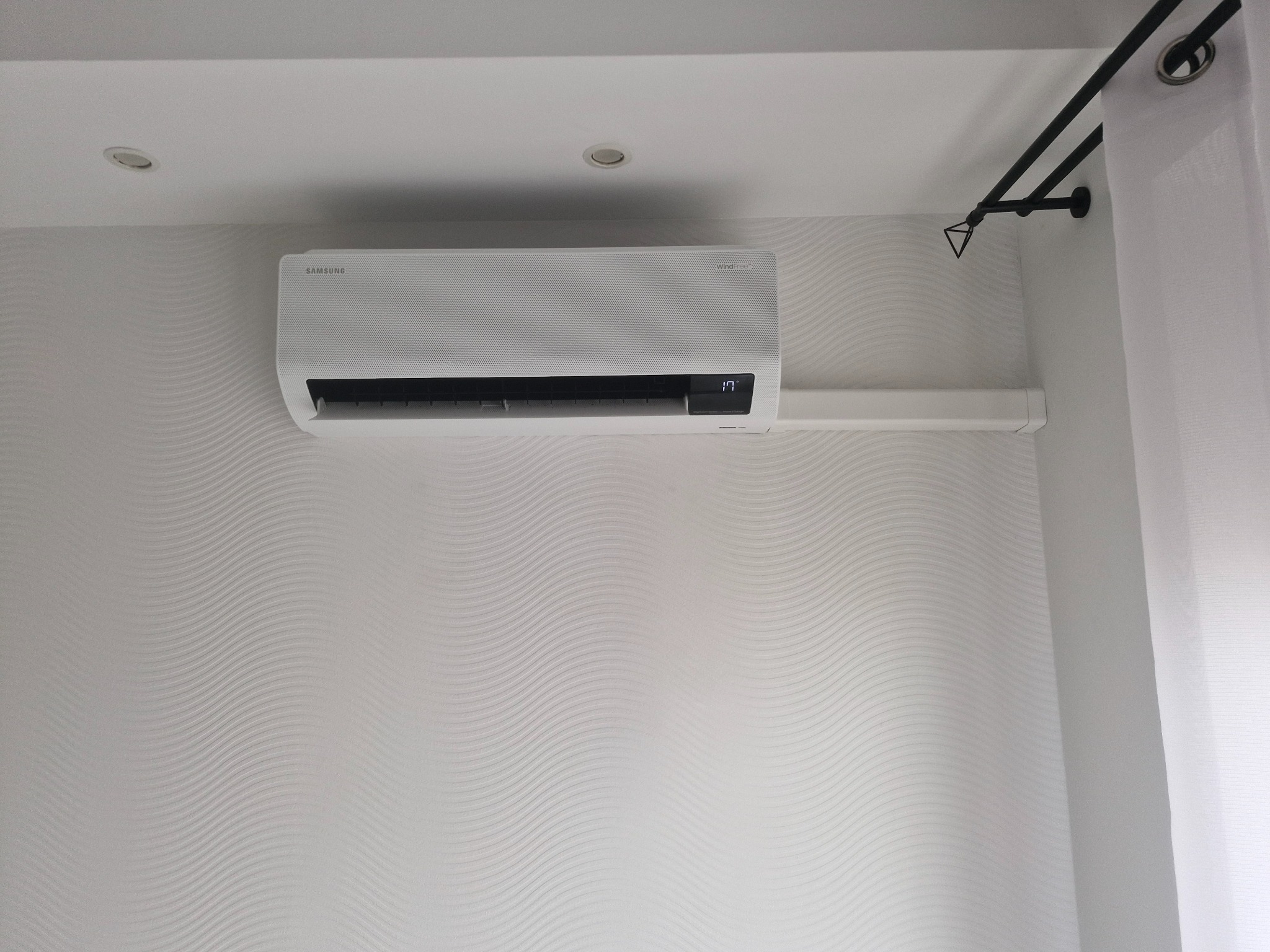 Biała klimatyzacja Samsung WindFree na ścianie z subtelnym wzorem fal, widoczny fragment karnisza i zasłony. Wyświetlacz pokazuje 17 stopni Celsjusza.