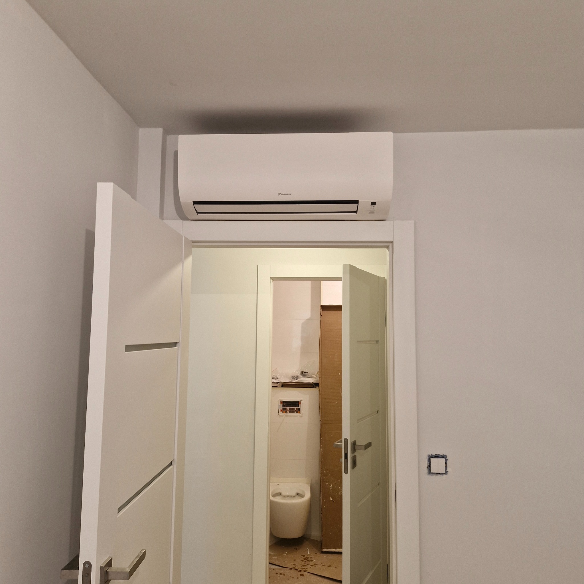 Biały klimatyzator Daikin zamontowany nad otwartymi, białymi drzwiami w trakcie remontu łazienki. Widoczne instalacje i toaleta w remoncie.
