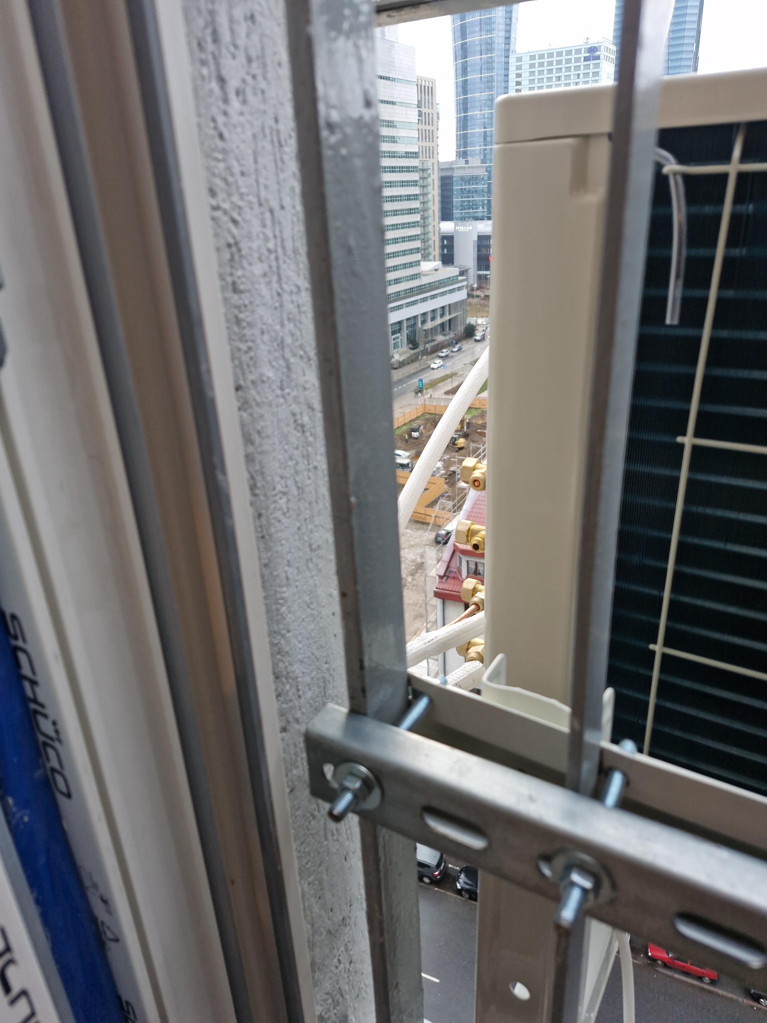 Zewnętrzna jednostka klimatyzacji zamontowana na balkonie z widokiem na miasto. Widoczne elementy montażowe i fragment okna.