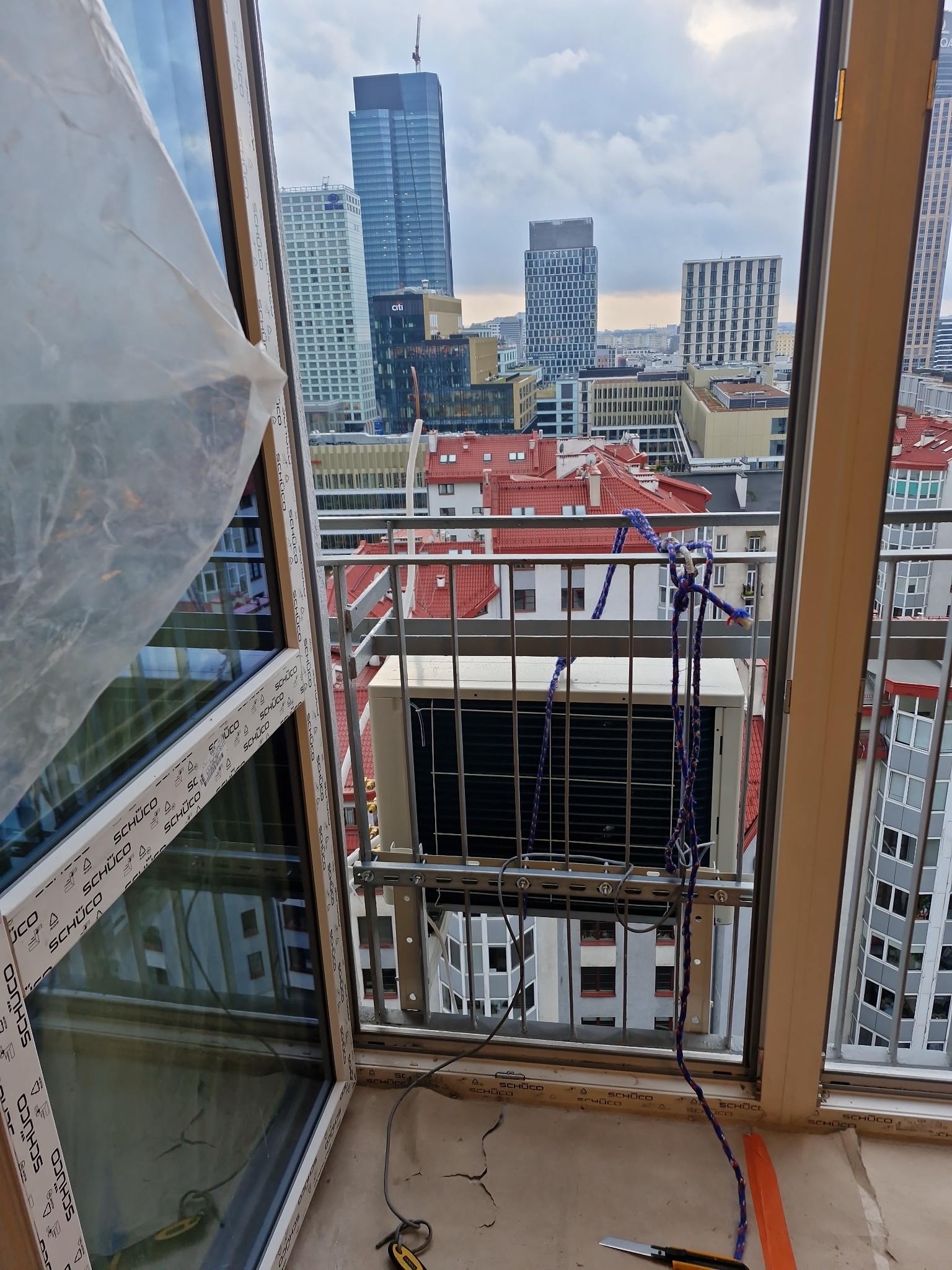 Montaż klimatyzatora na balkonie z widokiem na panoramę miasta. Urządzenie zabezpieczone linami, widoczne narzędzia. Okna marki Schüco.