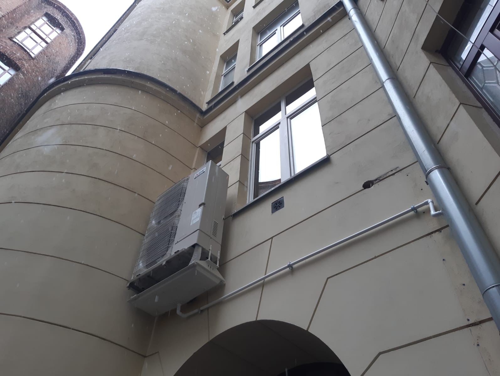 Klimatyzator zamontowany na elewacji budynku z widocznymi oknami i rurą spustową. Ujęcie z dołu podkreśla architekturę i instalację klimatyzacyjną.