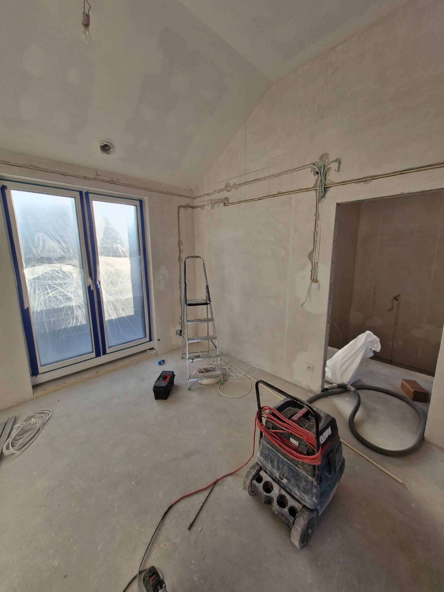 Wnętrze w stanie surowym z widoczną instalacją elektryczną na ścianach, drabiną, oknem zabezpieczonym folią i odkurzaczem przemysłowym.