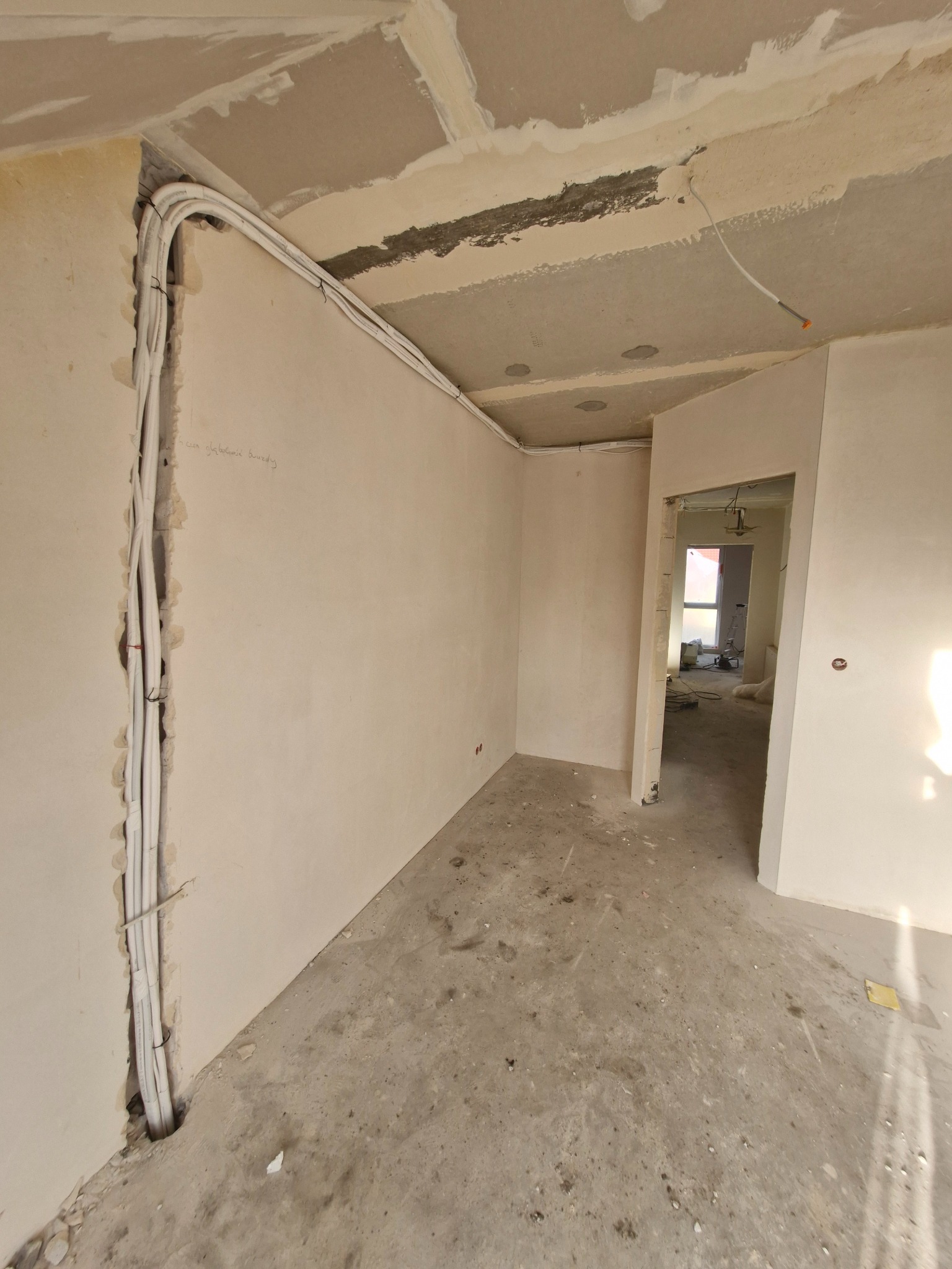 Wnętrze w trakcie remontu: białe kable instalacji elektrycznej prowadzone w bruzdach ściany, surowy sufit i posadzka. Widok na sąsiednie pomieszczenie.