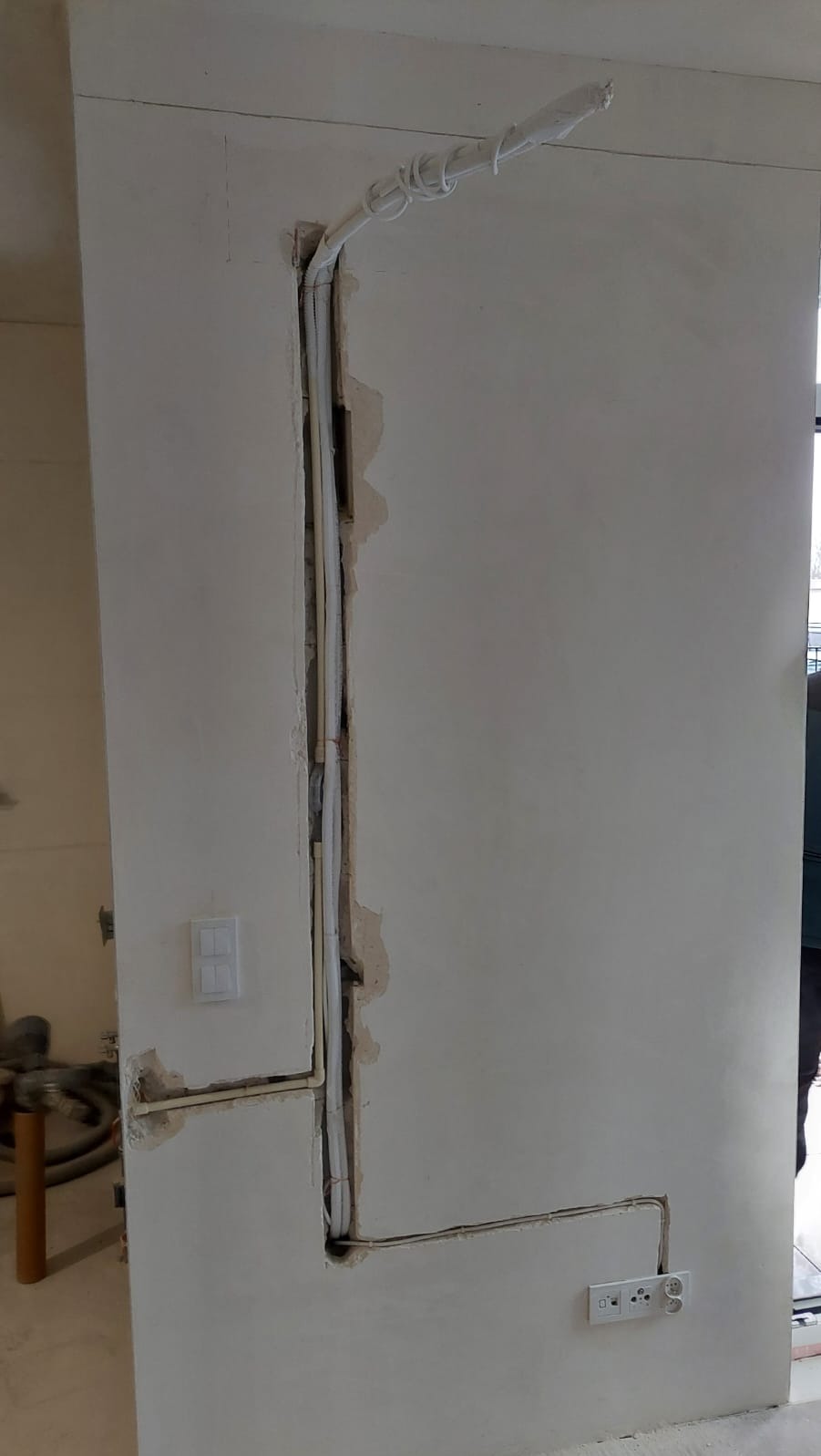 Biała ściana z wyżłobionym pionowym kanałem, w którym umieszczono rury i przewody. Widoczne gniazdka i włącznik. Etap prac instalacyjnych przed wykończeniem.