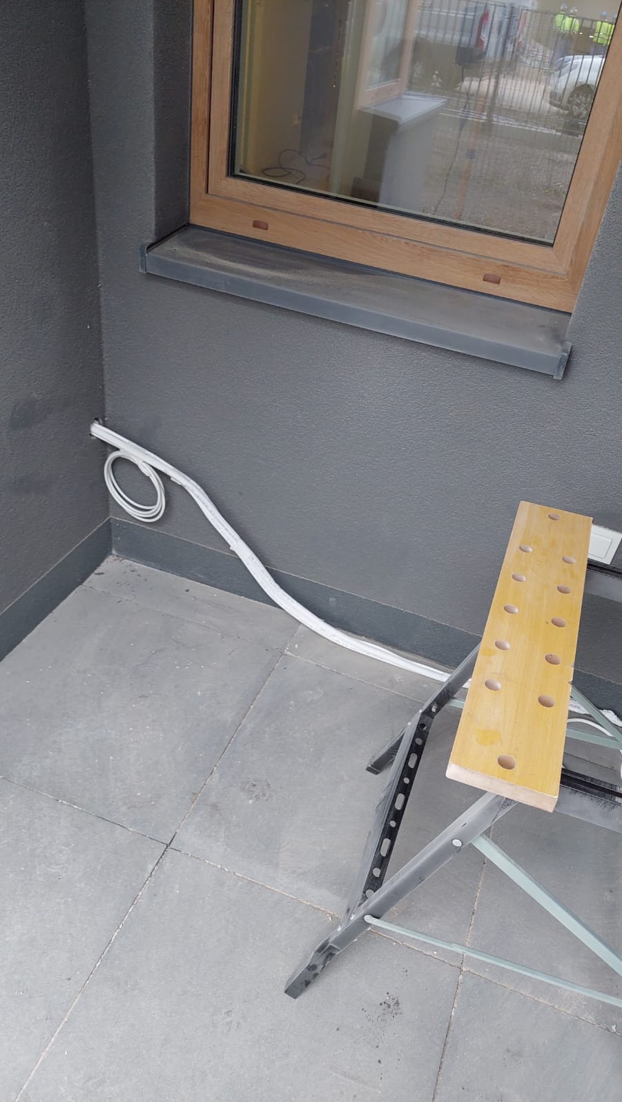 Szara ściana z oknem, obok biała listwa instalacyjna i rozłożony stół roboczy. Instalacja elektryczna lub klimatyzacyjna w trakcie montażu.