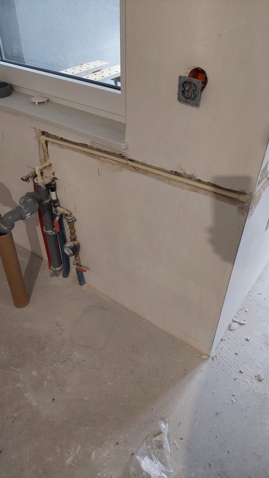 Instalacja wodna i elektryczna przy oknie: rury, zawory, licznik i gniazdko w ścianie. Surowy stan, przed wykończeniem. Widoczne niedoskonałości na ścianie.