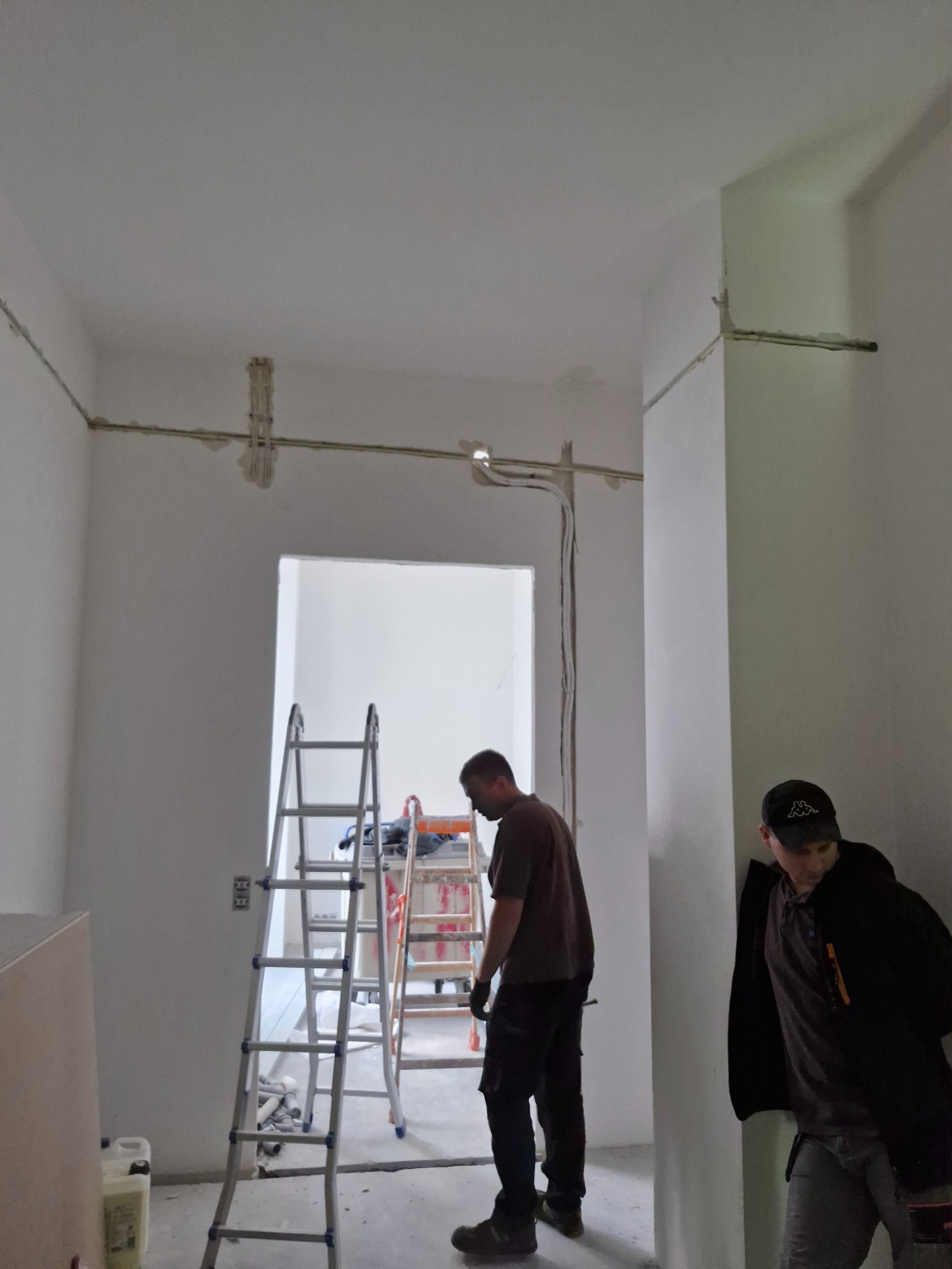 Instalacja elektryczna w surowym wnętrzu: widoczne przewody na ścianach, drabiny i dwóch mężczyzn w trakcie pracy. Realizacja w trakcie montażu.