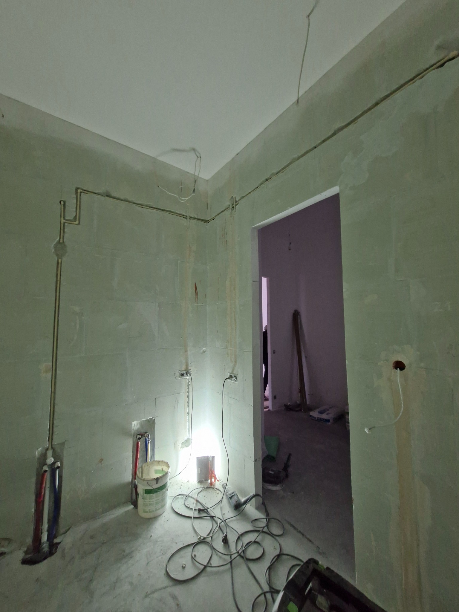 Surowy stan wnętrza: instalacja elektryczna i hydrauliczna w ścianach, przewody na podłodze, przygotowanie do wykończenia. Widok na sąsiednie pomieszczenie.