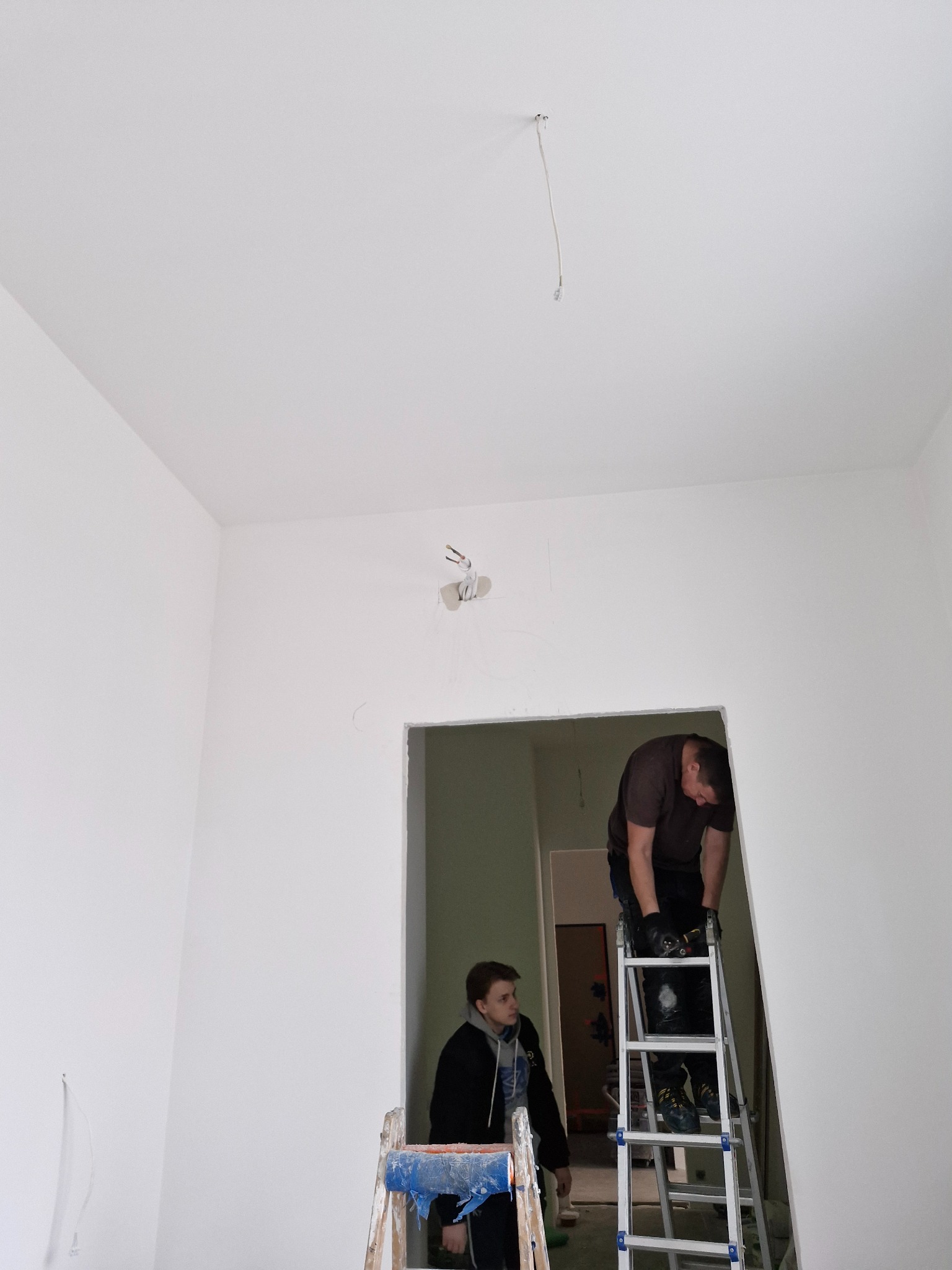 Montaż instalacji elektrycznej: elektryk na drabinie, w tle drugi pracownik. Przewody wystające z sufitu. Świeżo pomalowane ściany w jasnym pokoju.