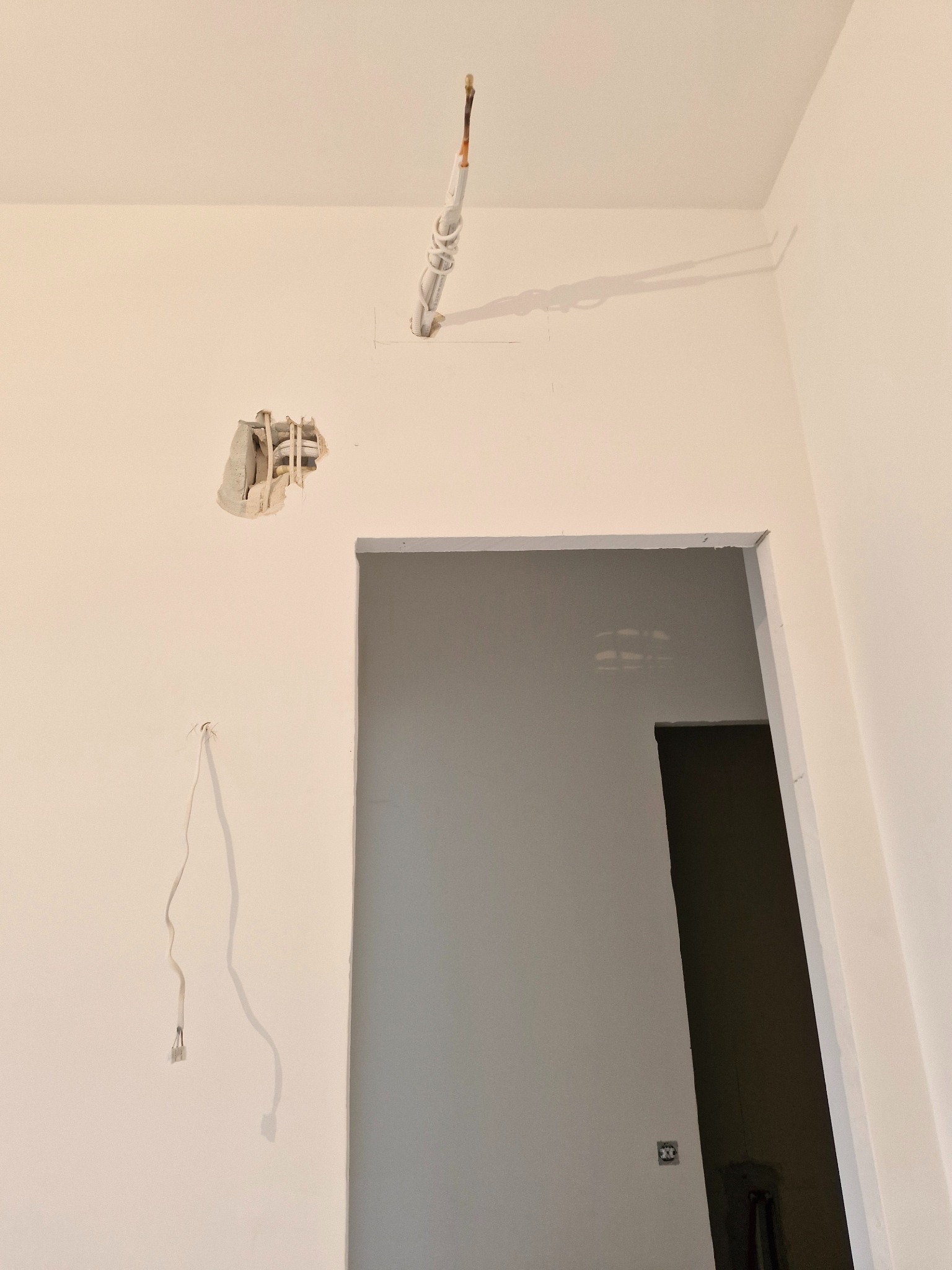 Niedokończona instalacja elektryczna w ścianie, widoczne kable i otwory. Prace remontowe w jasnym pomieszczeniu z otwartym przejściem.