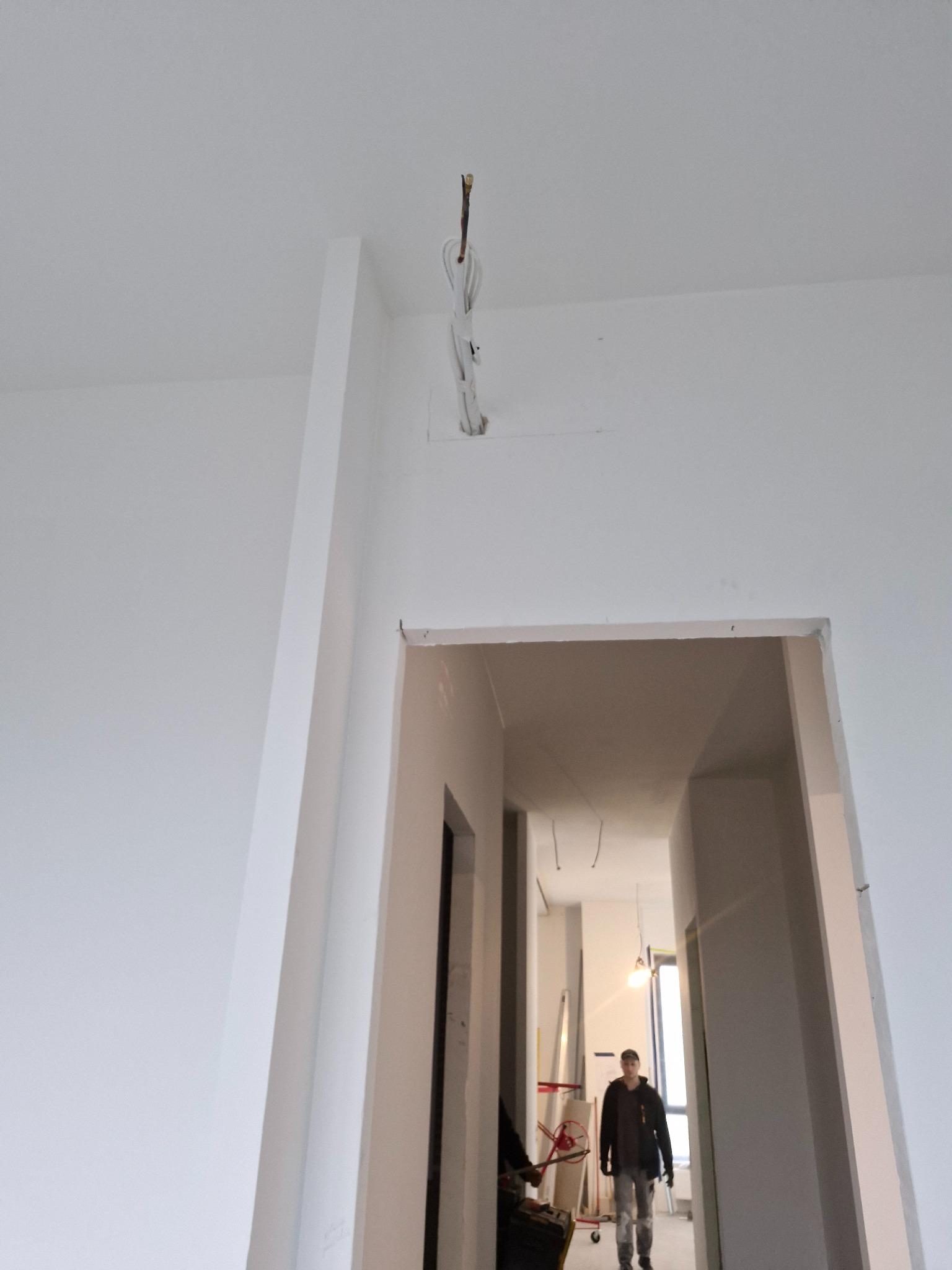 Wnętrze w trakcie remontu: białe ściany, wystające przewody elektryczne i sylwetka mężczyzny w oddali, sugerujące etap instalacji elektrycznej.
