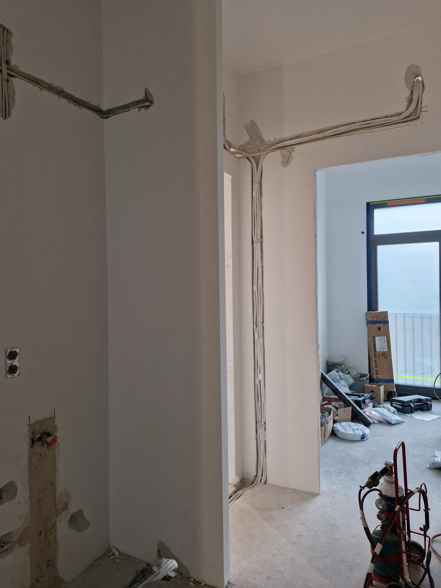 Instalacja przewodów wentylacyjnych/klimatyzacyjnych w surowym wnętrzu. Widoczne przewody przytwierdzone do ściany, narzędzia pracy w tle.