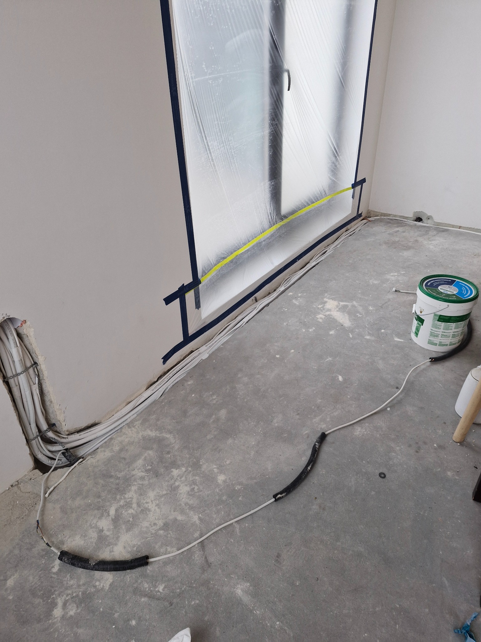 Instalacja przewodów w ścianie i na podłodze podczas remontu. Okno zabezpieczone folią. Widoczne kable, puszka z farbą i fragment drabiny.