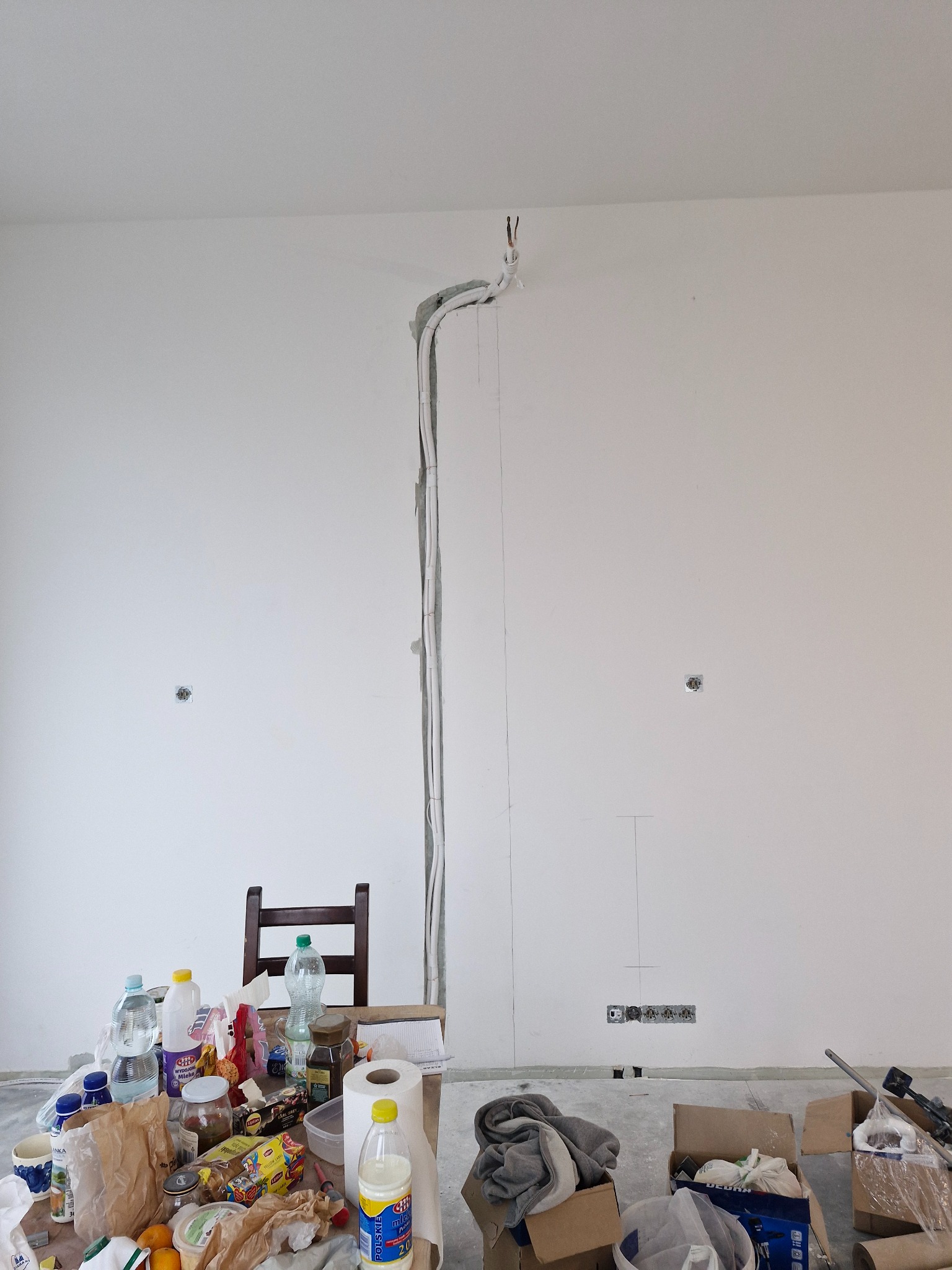 Biała ściana z wyżłobioną bruzdą na przewody, w tle stół z jedzeniem i narzędzia. Instalacja elektryczna w trakcie prac przygotowawczych.