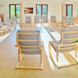 Jasne, przestronne wnętrze z krzesłami ustawionymi w okrąg, tworzącymi przestrzeń do spotkań grupowych; widok na zewnątrz przez okna, z obrazem na ścianie przedstawiającym krajobraz.