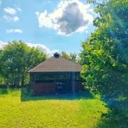 Drewniana altana z brązowym dachem, stolikiem i krzesłami w środku, otoczona zielonym trawnikiem i drzewami w słoneczny dzień.