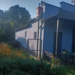 Jasnobłękitny dom jednorodzinny z czerwonym kominem, widoczny z boku, z zadbanym ogrodem i budą dla psa w tle, oświetlony porannym słońcem.