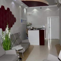 Jasne wnętrze poczekalni z szarą sofą, stolikiem kawowym i recepcją, akcenty bordowe, logo 'Dent Kraków' na ścianie.