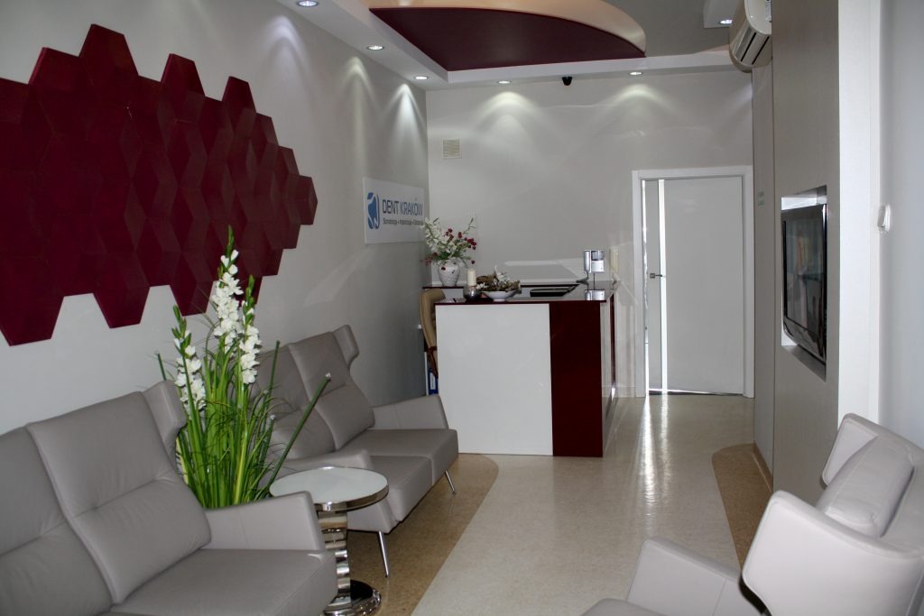 Jasne wnętrze poczekalni z szarą sofą, stolikiem kawowym i recepcją, akcenty bordowe, logo 'Dent Kraków' na ścianie.