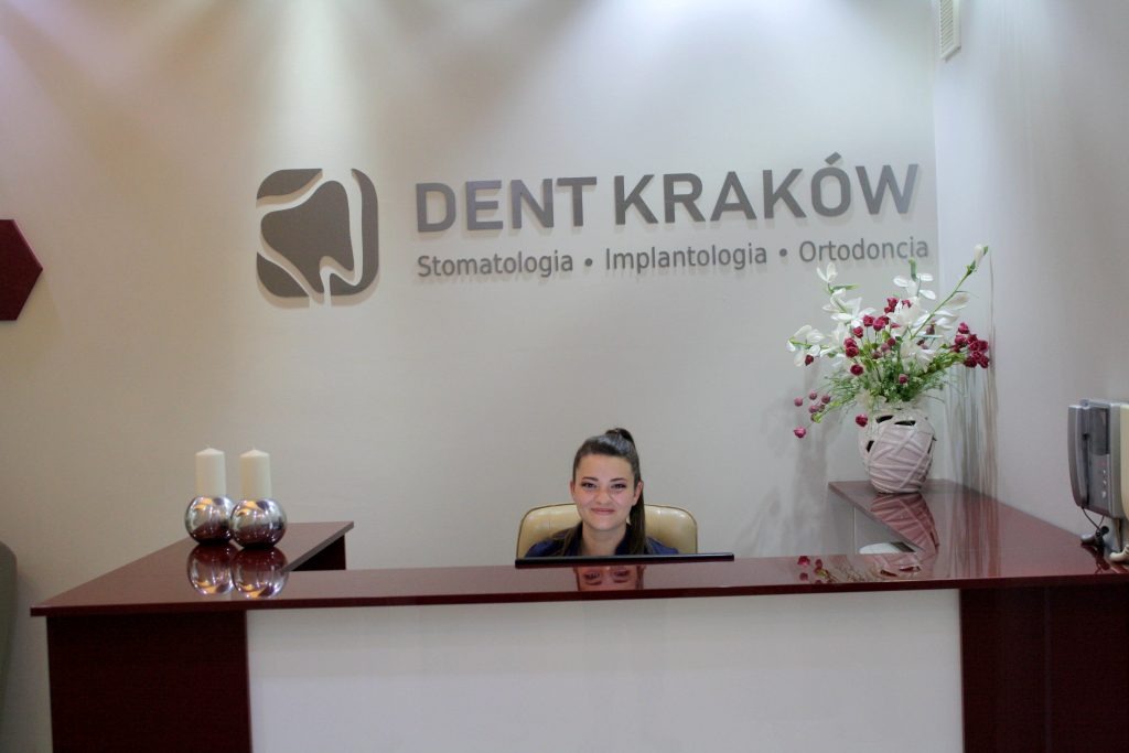 Recepcjonistka w recepcji kliniki stomatologicznej Dent Kraków, z logo firmy na ścianie, dekoracyjnymi świecami i kwiatami w tle.