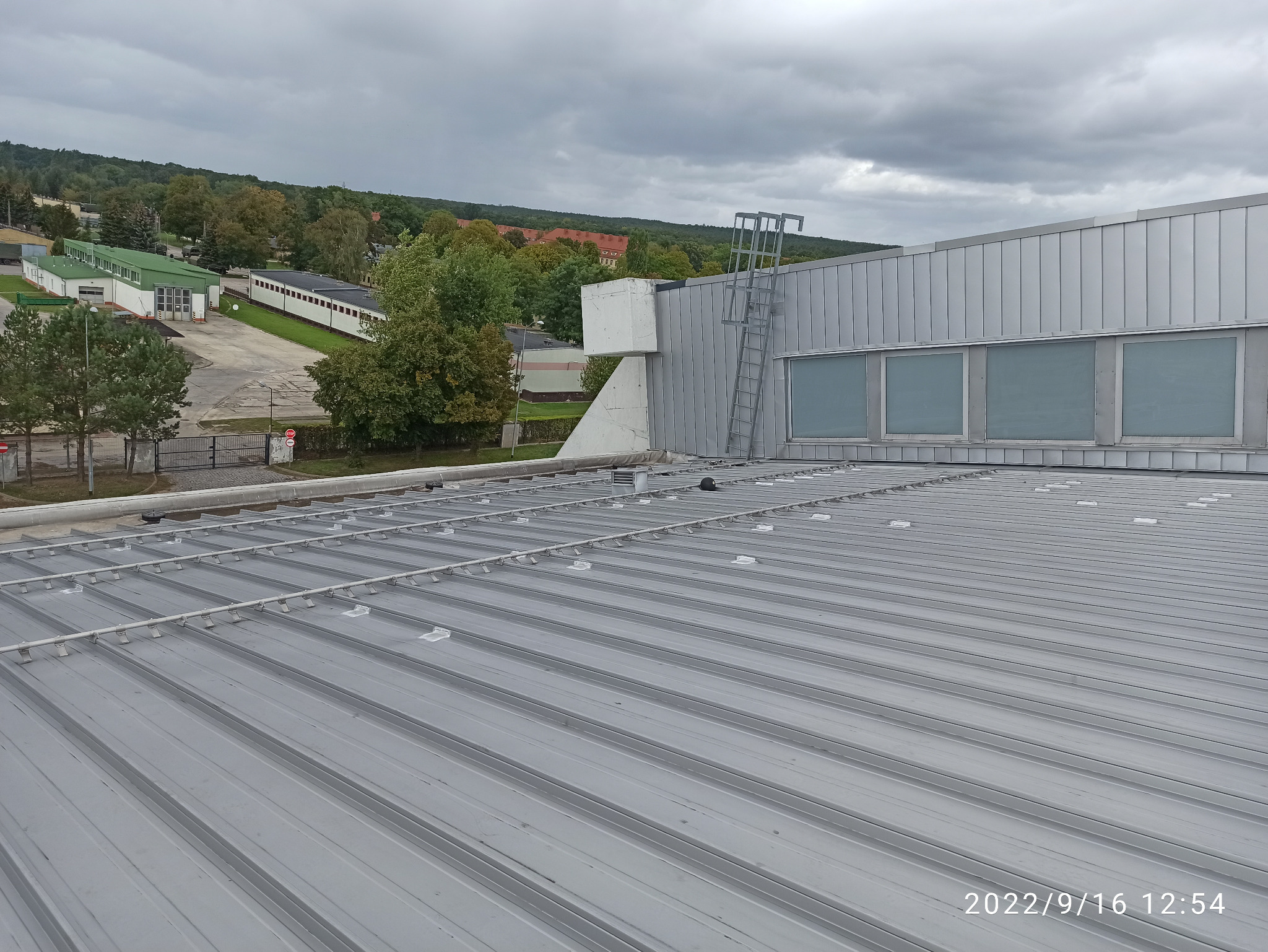 Szary, blaszany dach przemysłowy z widocznymi mocowaniami i konstrukcją wsporczą, w tle budynki i drzewa pod pochmurnym niebem.