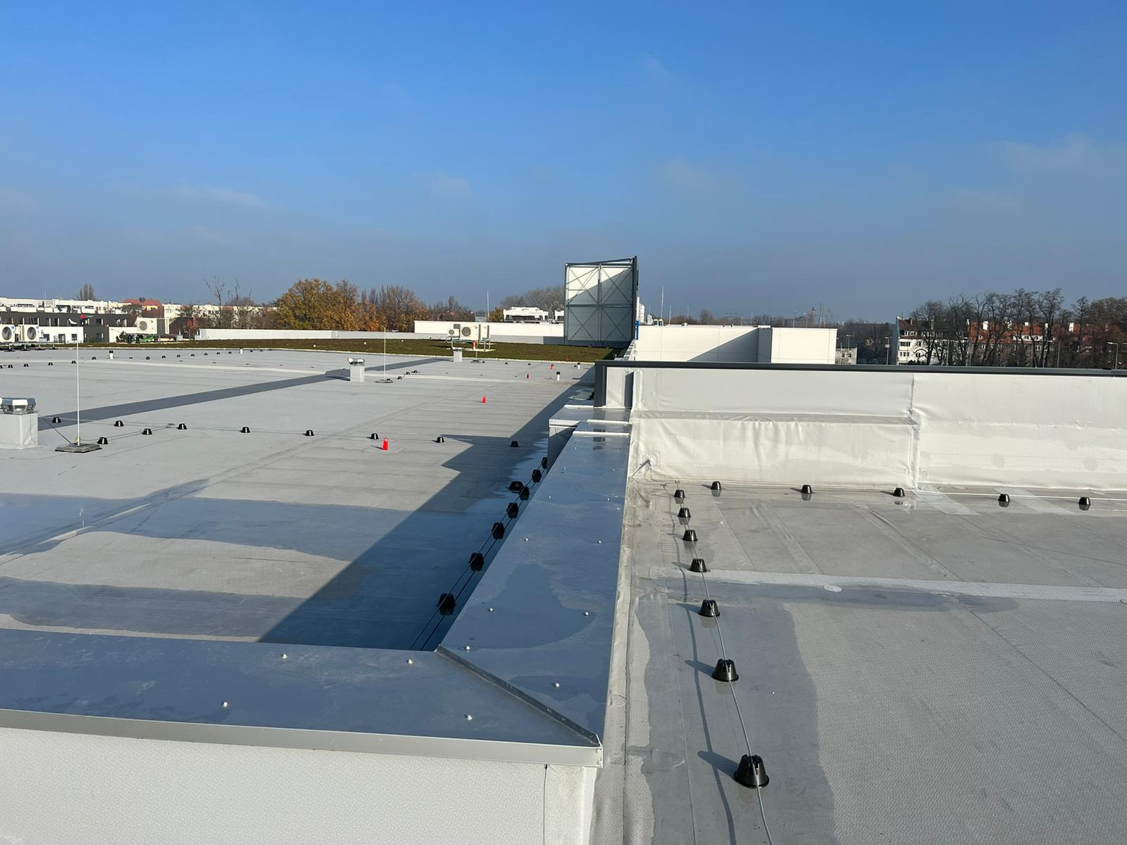 Widok z góry na płaski dach pokryty jasną membraną, z widocznymi elementami mocującymi i systemem odwadniającym, w tle budynki i drzewa pod błękitnym niebem.