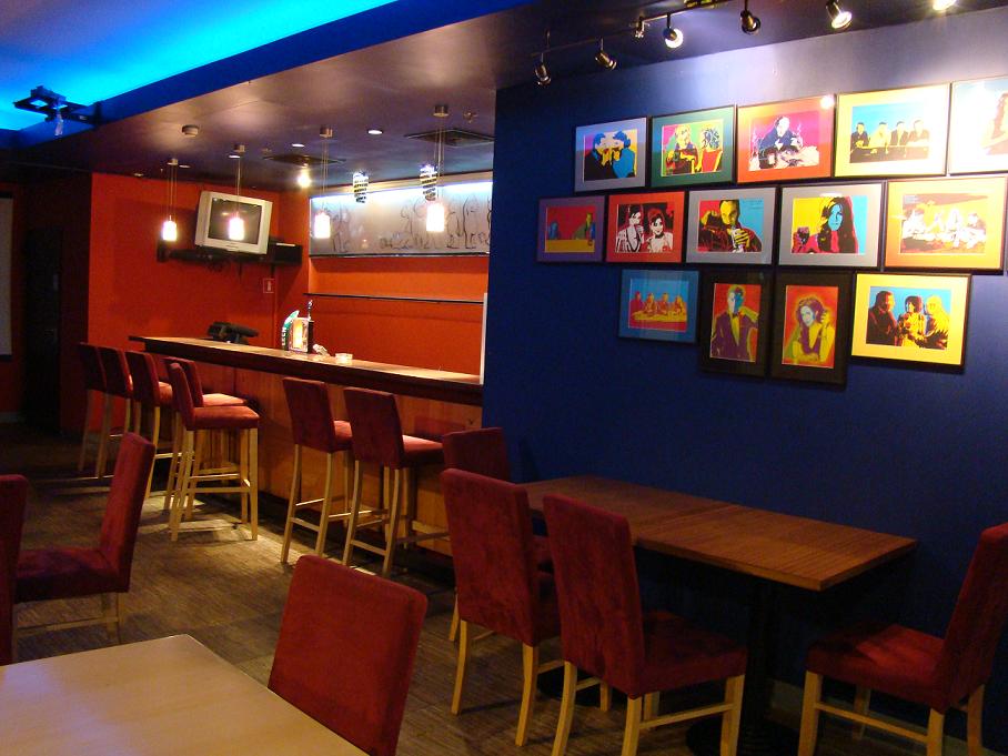 Wnętrze sali konferencyjnej z barem, krzesłami barowymi i stołami dla uczestników, ściana dekorowana serią oprawionych grafik w stylu pop-art.