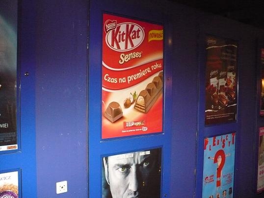 Rząd plakatów w kinie, w tym reklama batonów KitKat Senses z hasłem 'Czas na premierę roku' i plakaty filmowe, umieszczone na niebieskiej ścianie.