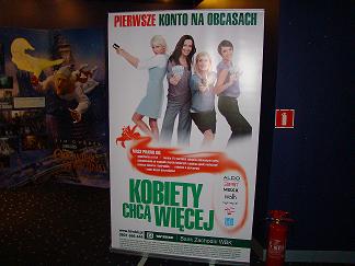 Plakat reklamowy filmu 'Kobiety chcą więcej' w kinie, widoczny obok gaśnica i fragment innego plakatu z Shrekiem.