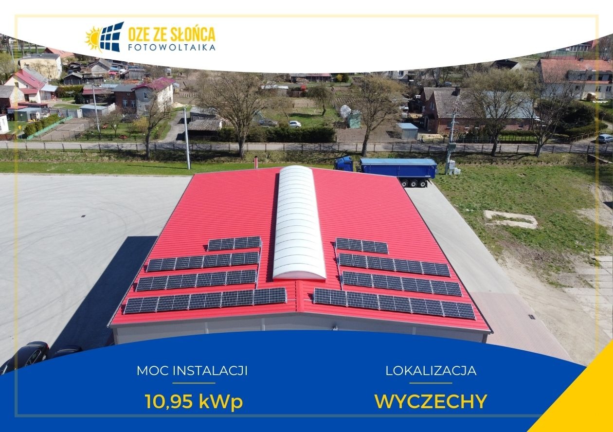 Widok z góry na czerwoną halę z zamontowanymi panelami słonecznymi, w tle zabudowa wiejska i niebieska ciężarówka.