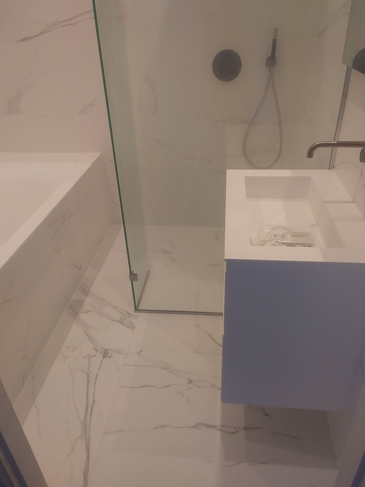 Nowoczesna łazienka wykończona białym marmurem, z kabiną prysznicową ze szklaną ścianką, umywalką z szafką w kolorze gołębim oraz baterią w odcieniu szczotkowanego brązu. Na umywalce widoczne...