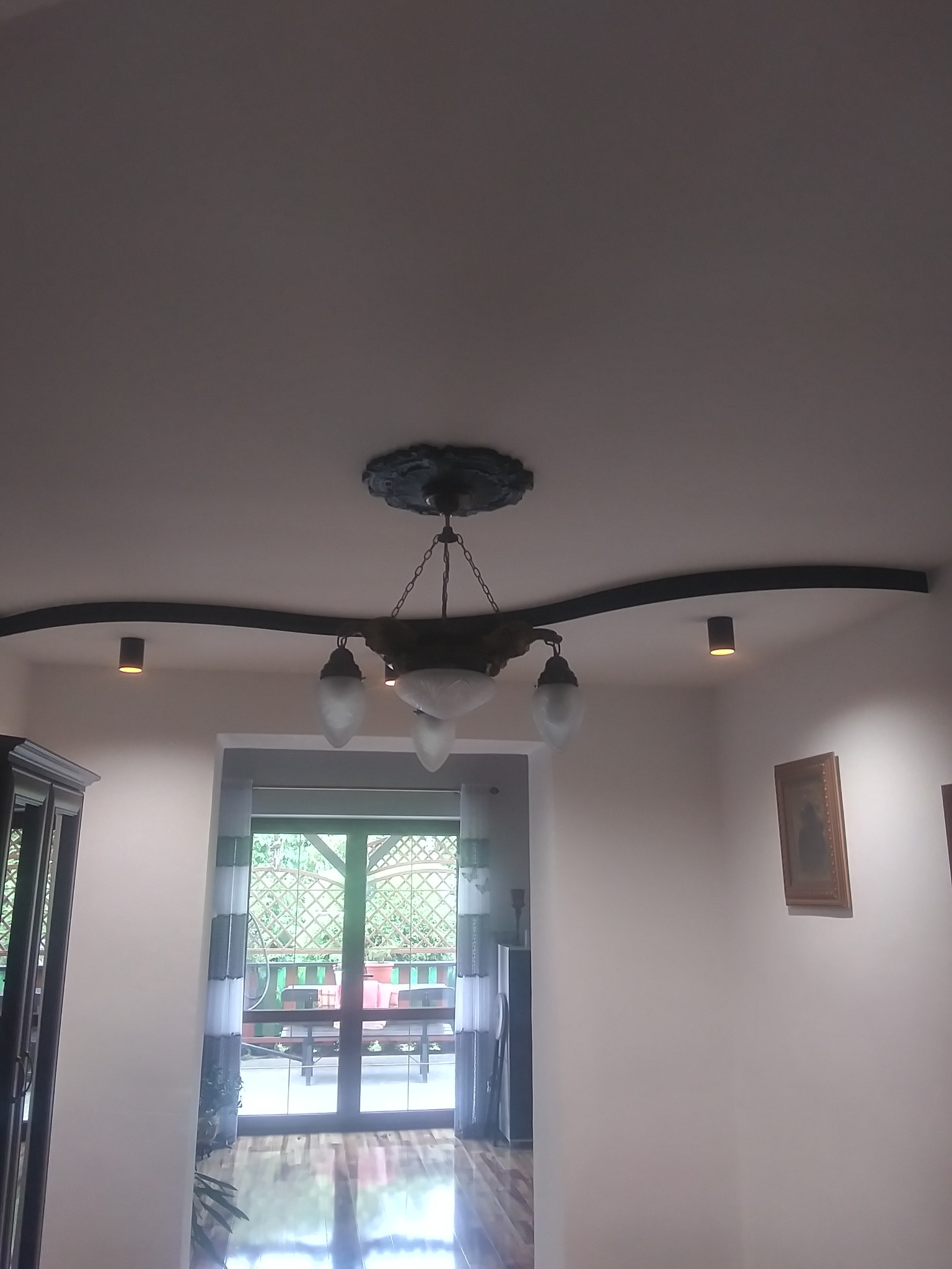 Fragment salonu z dekoracyjnym sufitem podwieszanym, oświetleniem punktowym i klasycznym żyrandolem z mlecznymi kloszami, widok na przeszklone drzwi balkonowe.