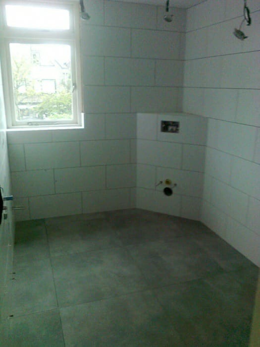 Wnętrze łazienki w trakcie układania płytek ściennych i podłogowych, widoczne przygotowane instalacje wodne i okno z widokiem na zieleń.