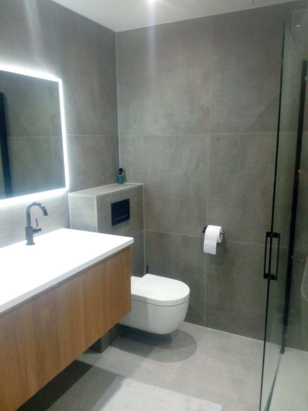 Nowoczesna łazienka z szarymi płytkami, podświetlanym lustrem, drewnianą szafką pod umywalką i kabiną prysznicową z czarnym uchwytem.