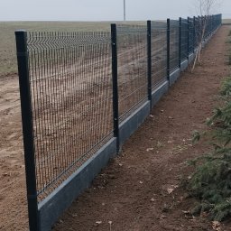 BTW Błażej Wrzesiński - Nowoczesne ogrodzenie panelowe z podmurówką w Krogulcu. Metalowa siatka, betonowa podstawa, schludny montaż. Widok na pole i słup energetyczny w tle.