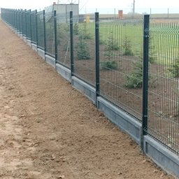BTW Błażej Wrzesiński - Długi, panelowy, metalowy płot z podmurówką, ciągnący się wzdłuż ziemistej działki. Widoczne słupki i siatka ogrodzeniowa. W tle zieleń i budynki.