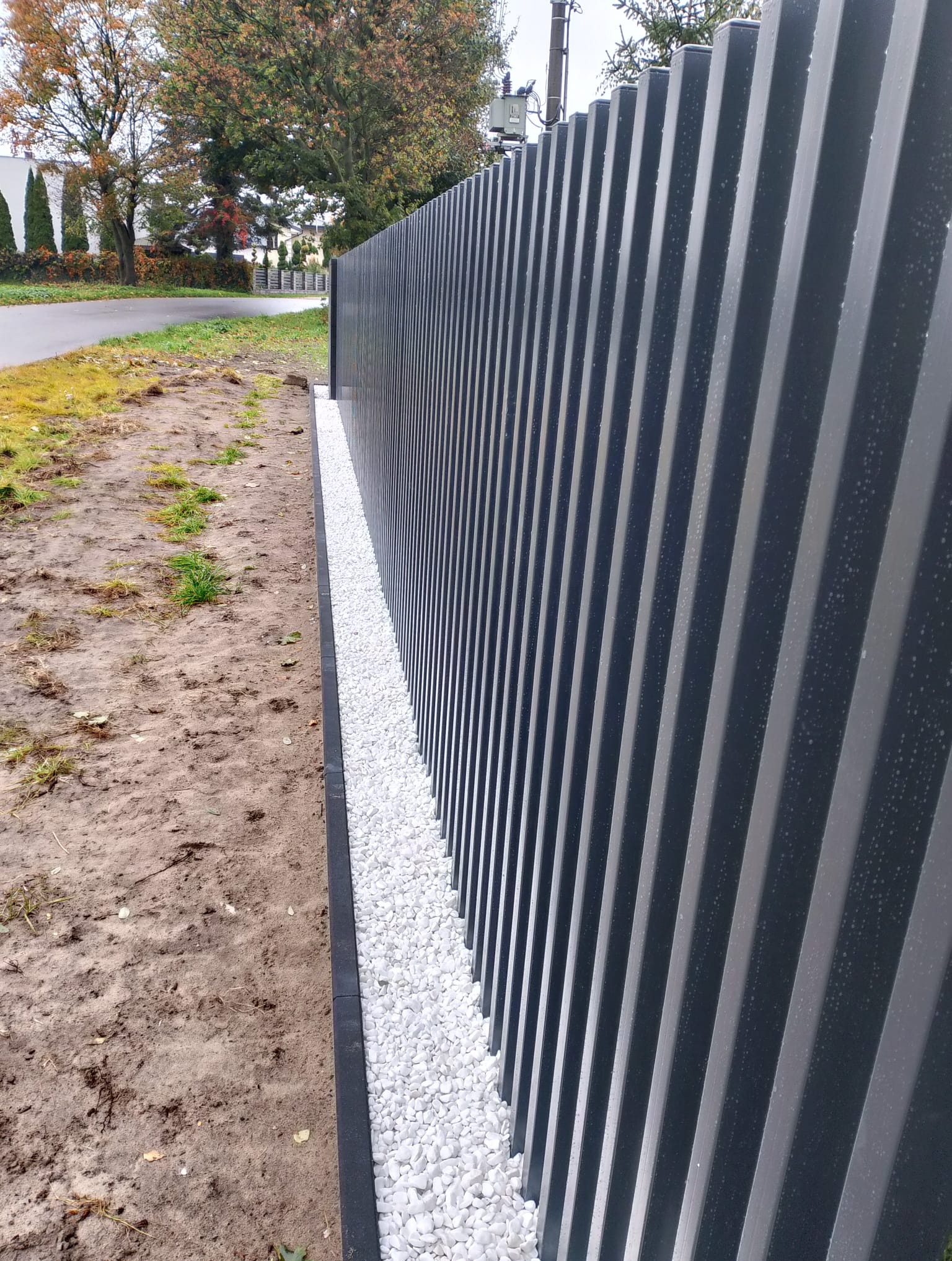 Nowoczesne, grafitowe ogrodzenie panelowe z dekoracyjnym podsypem z białych kamieni. Widok z boku, podkreślający rytmiczną strukturę paneli.