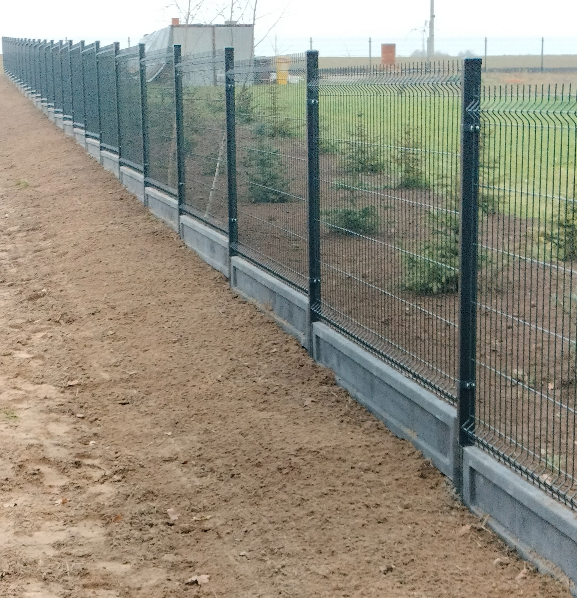 Długi, panelowy, metalowy płot z podmurówką, ciągnący się wzdłuż ziemistej działki. Widoczne słupki i siatka ogrodzeniowa. W tle zieleń i budynki.