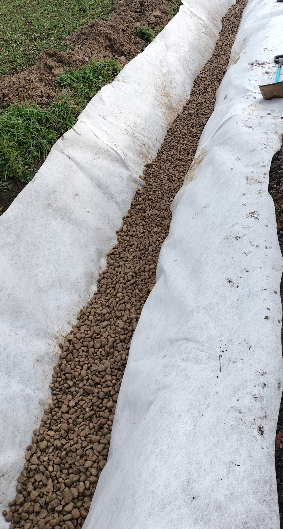 Drenaż liniowy w trakcie budowy: wykop wyłożony białą geowłókniną, wypełniony brązowym żwirem drenażowym. Widok z góry, fragment narzędzia w prawym górnym rogu.