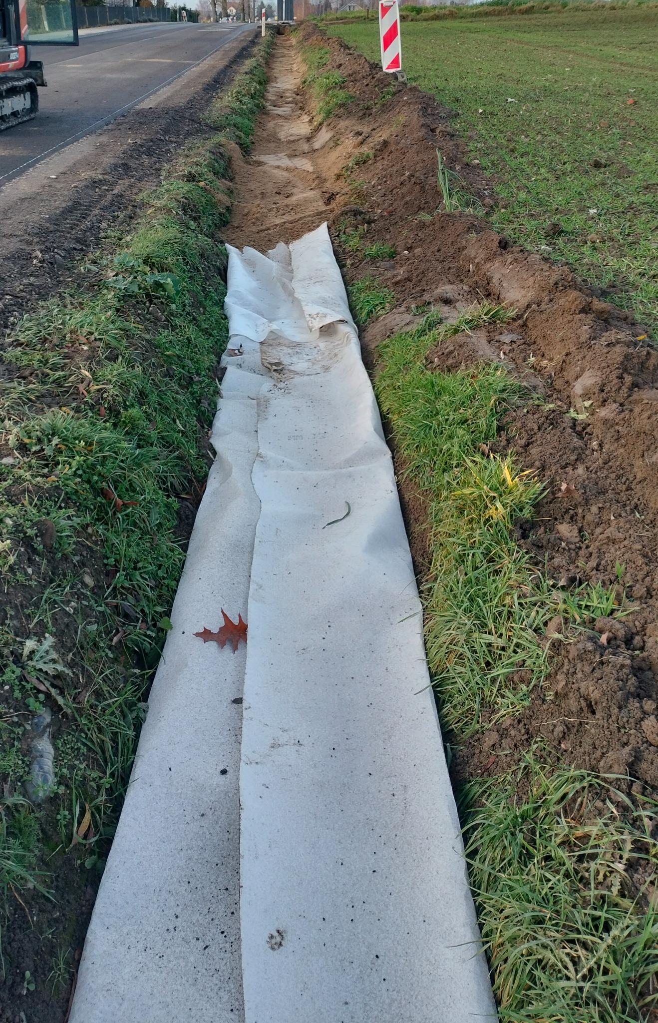 Wykopany rów wyłożony białą geowłókniną, widoczny fragment koparki i znak ostrzegawczy. Przygotowanie pod drenaż na gruncie z trawą i ziemią.